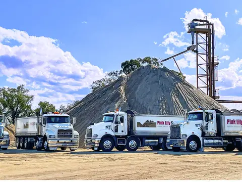 Pink Lily Sands trucks parked in front of a large sand stockpile and processing plant at a Heidelberg Materials Australia quarry operation under blue skies.
