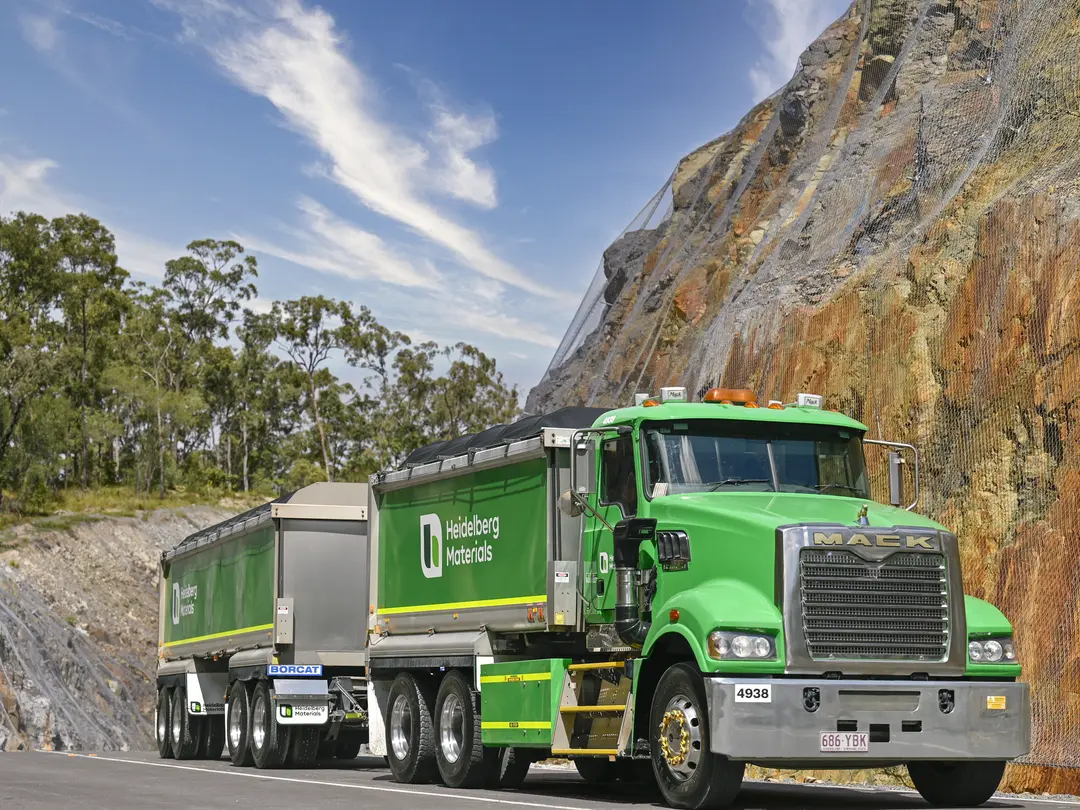 A green Heidelberg Materials Mack truck with trailer driving through a quarry haul road beside a steep rock face fitted with protective mesh under a blue sky.