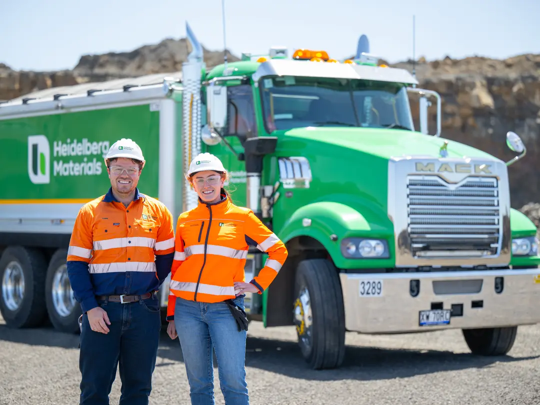 Two Heidelberg Materials employees wearing PPE and hard hats standing in front of a green Heidelberg Materials haul truck at a quarry site.
