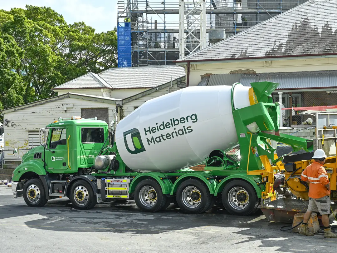 A green ready mix concrete truck with "Heidelberg Materials" branding on its drum is parked at a construction site. Workers in helmets and high-visibility clothing stand near the truck's rear, while buildings and scaffolding are visible in the background.